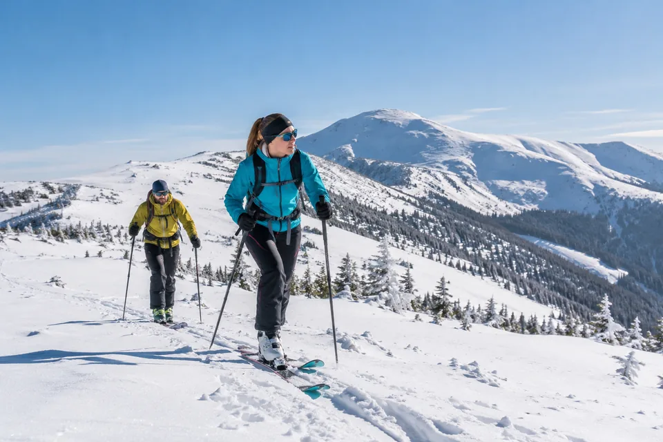 Dva skialpinisté stoupají na lyžích zasněženým svahem.